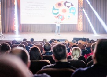 Audience attentively watching a presentation in a conference setting, with stage lights and speaker visible.