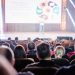 Audience attentively watching a presentation in a conference setting, with stage lights and speaker visible.