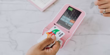 Hand using a contactless card on a pink POS terminal for a wireless transaction.