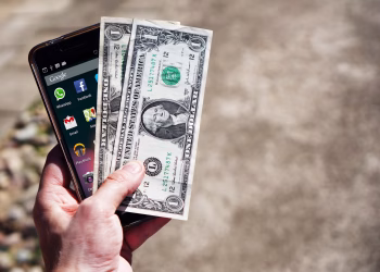 Close-up of a hand holding US dollar bills and a smartphone outdoors, showcasing financial technology.