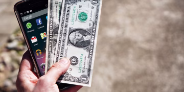Close-up of a hand holding US dollar bills and a smartphone outdoors, showcasing financial technology.
