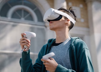 Low angle of young male in blue hoodie using modern wireless virtual reality headset and controllers near building on street