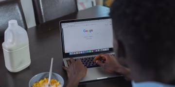 A person browsing Google on a laptop while having cereal breakfast at the table.