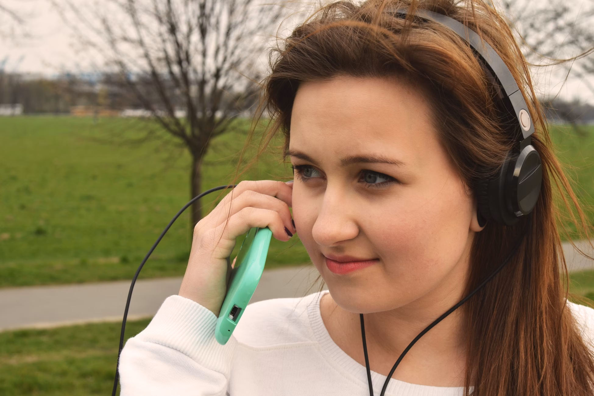 A young woman listens to music with headphones in a lush green park setting.