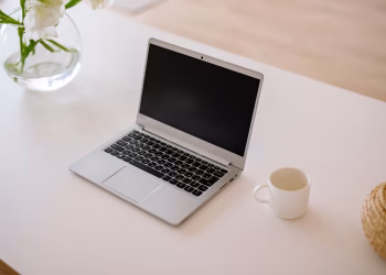 A minimalist workspace with an open laptop, coffee mug, and vase on a bright indoor table.