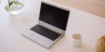 A minimalist workspace with an open laptop, coffee mug, and vase on a bright indoor table.