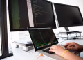 A developer writes code on a laptop in front of multiple monitors in an office setting.