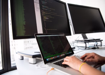 A developer writes code on a laptop in front of multiple monitors in an office setting.