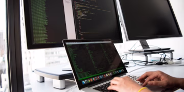 A developer writes code on a laptop in front of multiple monitors in an office setting.