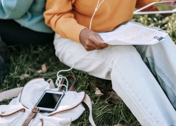 High angle of unrecognizable students in casual outfit sitting on lawn in park doing homework and using tablet