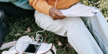 High angle of unrecognizable students in casual outfit sitting on lawn in park doing homework and using tablet