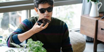 A man wearing sunglasses uses a smartphone's voice assistant in a cozy indoor setting.