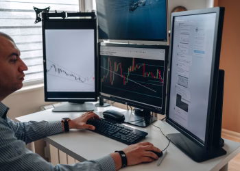 A professional in an office analyzing financial charts on multiple monitors, using advanced technology.