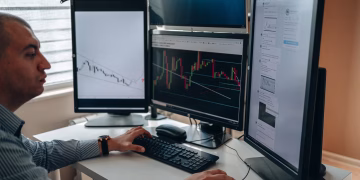 A professional in an office analyzing financial charts on multiple monitors, using advanced technology.