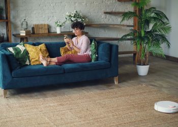 Woman relaxing on a stylish blue sofa at home, using a smartphone with a robotic vacuum on the floor.