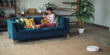 Woman relaxing on a stylish blue sofa at home, using a smartphone with a robotic vacuum on the floor.