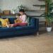 Woman relaxing on a stylish blue sofa at home, using a smartphone with a robotic vacuum on the floor.