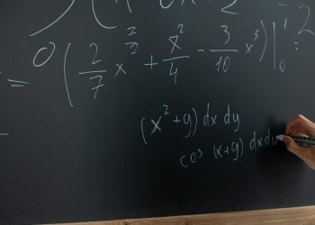 Hand writing mathematical equations on a chalkboard in a classroom setting.