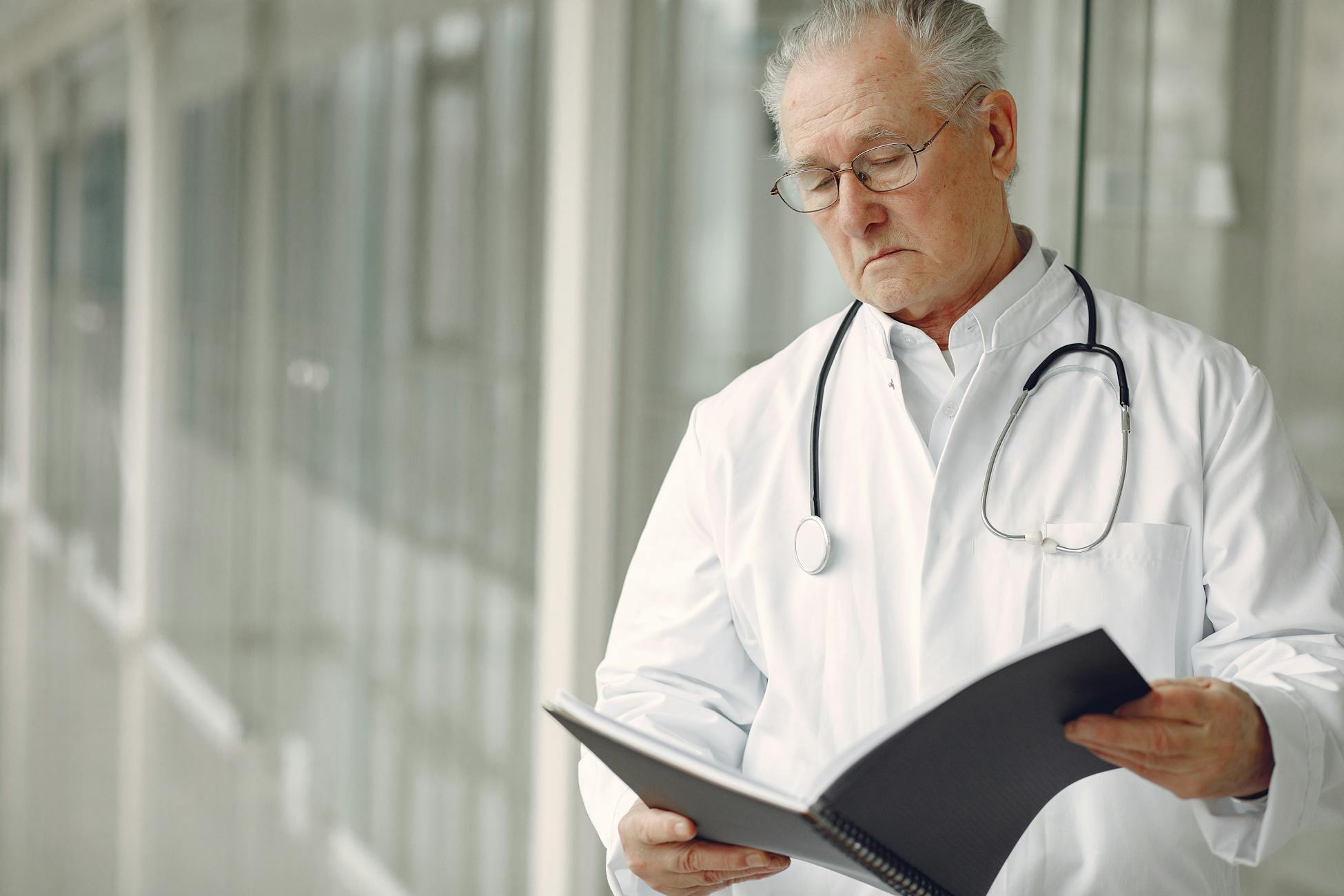 Crop concentrated professional senior male physician in white robe with stethoscope standing in modern hospital hallway and reading medical case records in daylight