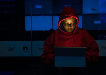 A woman in red hoodie works intensely on her laptop indoors, illuminated by screen light.