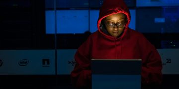 A woman in red hoodie works intensely on her laptop indoors, illuminated by screen light.