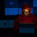 A woman in red hoodie works intensely on her laptop indoors, illuminated by screen light.