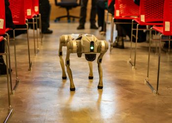 A robotic dog navigates an indoor setting amidst red chairs, showcasing technology in modern environments.