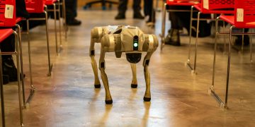 A robotic dog navigates an indoor setting amidst red chairs, showcasing technology in modern environments.
