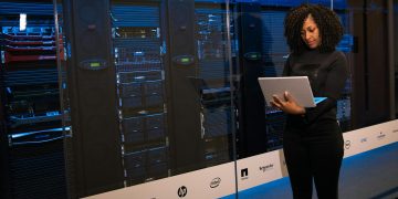 A female engineer using a laptop while monitoring data servers in a modern server room.