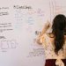 Female student writing circuit diagrams and logic gates in an educational setting on a whiteboard.