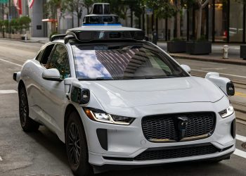 A white autonomous vehicle navigating a city street, reflecting urban architecture in daylight.