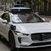 A white autonomous vehicle navigating a city street, reflecting urban architecture in daylight.