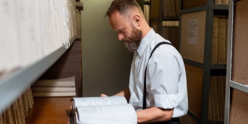 Bearded man in suspenders examining large book in archive room with wooden shelves.
