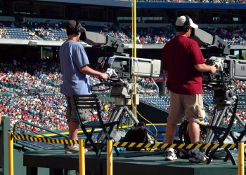 Camera operators capturing a live sports event at a crowded outdoor stadium filled with spectators.