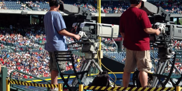 Camera operators capturing a live sports event at a crowded outdoor stadium filled with spectators.