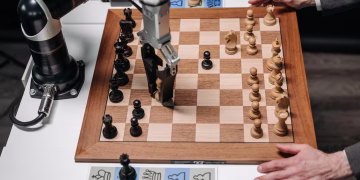 A robotic arm playing chess against a human hand on an electronic chessboard indoors.