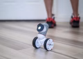 A close-up of a modern wheeled robot on a wooden floor with legs in the background.