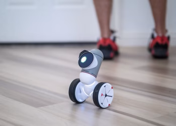 A close-up of a modern wheeled robot on a wooden floor with legs in the background.
