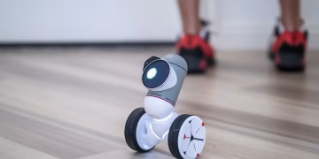 A close-up of a modern wheeled robot on a wooden floor with legs in the background.