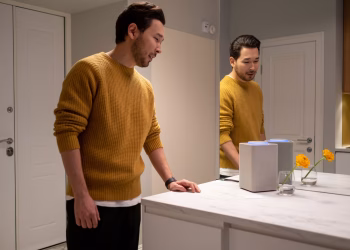 A man in a sweater reflects in a mirror next to a smart speaker in a modern home setting.
