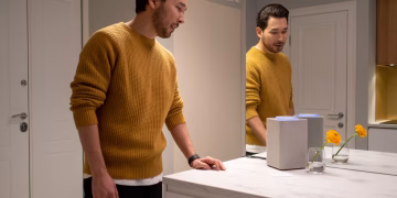 A man in a sweater reflects in a mirror next to a smart speaker in a modern home setting.