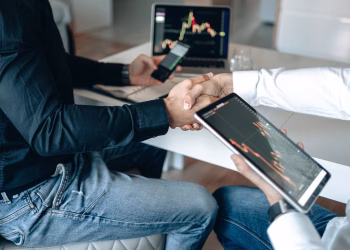 Two business professionals shaking hands with financial graphs on digital devices.