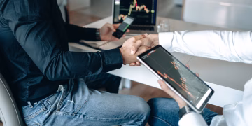 Two business professionals shaking hands with financial graphs on digital devices.