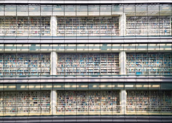 Sunlit view of a modern library facade with multiple levels of bookshelves behind glass walls.