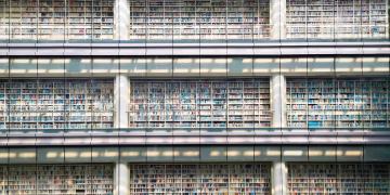 Sunlit view of a modern library facade with multiple levels of bookshelves behind glass walls.