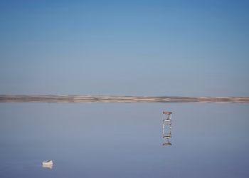 A solitary chair reflects on a calm salt flat under a clear blue sky, creating a serene scene.