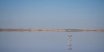 A solitary chair reflects on a calm salt flat under a clear blue sky, creating a serene scene.