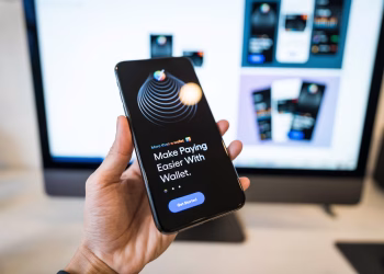 Hand holding smartphone displaying digital wallet app interface, blurred monitor in background.