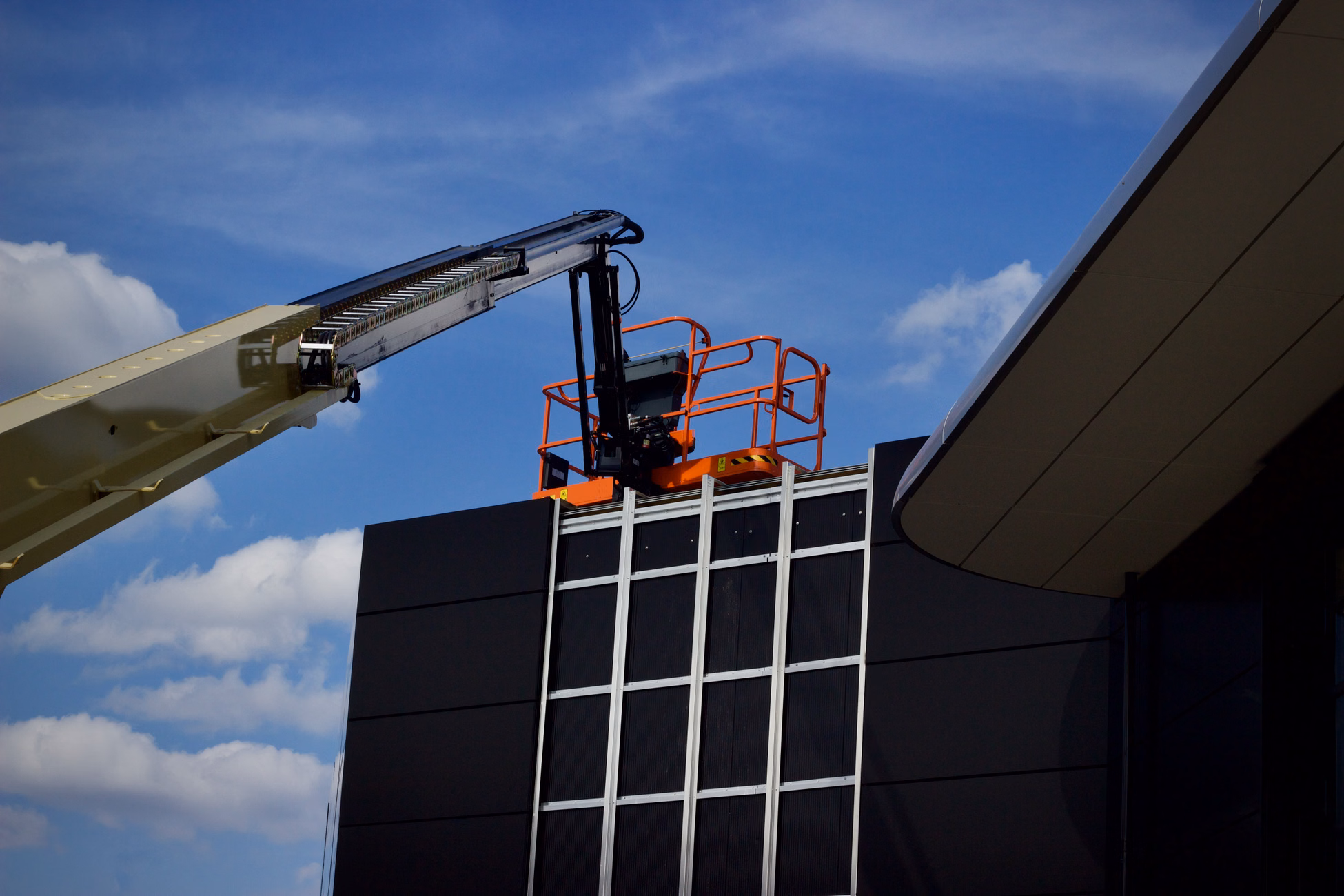 Aerial lift near modern building facade against a bright blue sky with scattered clouds.