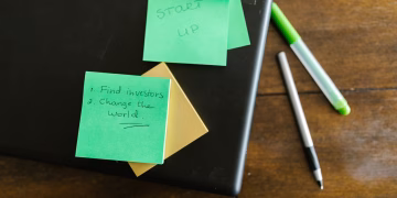Green sticky notes with startup goals on a wooden desk with pens.
